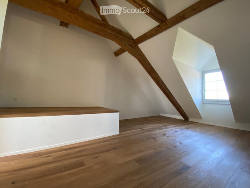 Empty attic space with wooden beams, wooden flooring, and a small window letting in natural light.