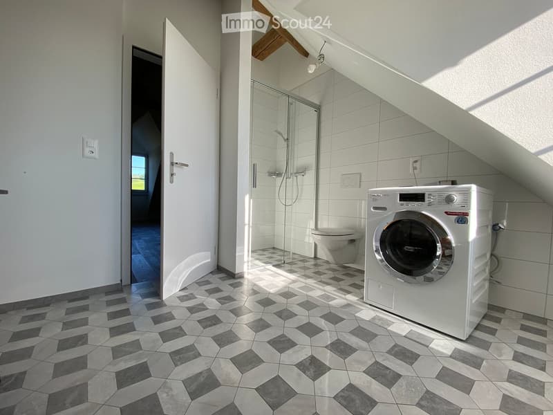 Modern bathroom with a washing machine, toilet, and glass-enclosed shower. The floor features a geometric tile pattern in gray and white.