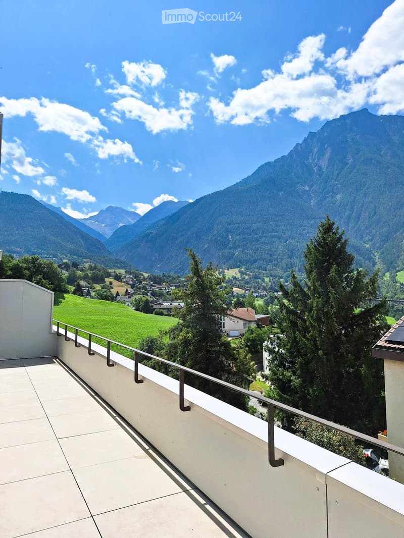 Balcony with a panoramic mountain view, surrounded by greenery and a small village below.