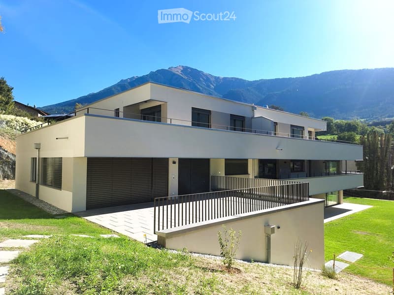 Modern two-story house with a flat roof, large windows, and a balcony overlooking a scenic mountain view. Features include a garage, raised ground floor, and a well-maintained lawn.