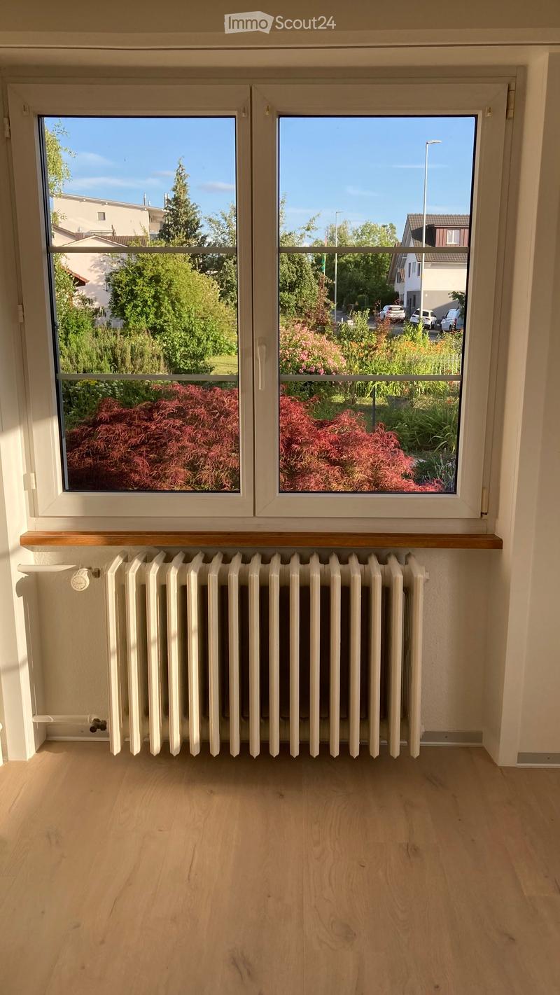 Empty room with wooden flooring, large windows showing a garden view, and a radiator below the windows.