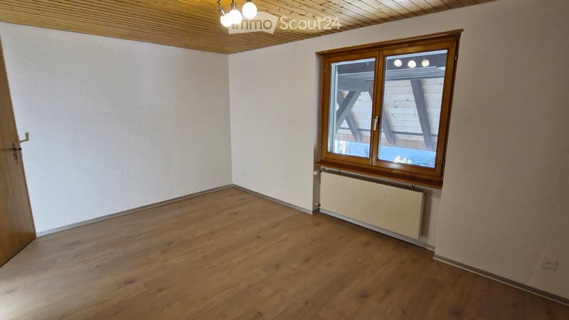 Empty room with wooden flooring, white walls, and a large window with wooden frame. The ceiling has wooden paneling and built-in lighting.