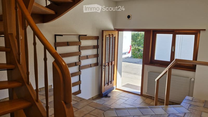 Spacious hallway with wooden staircase, tiled floor, and open doorway leading outside. Features a coat rack and window with blinds.