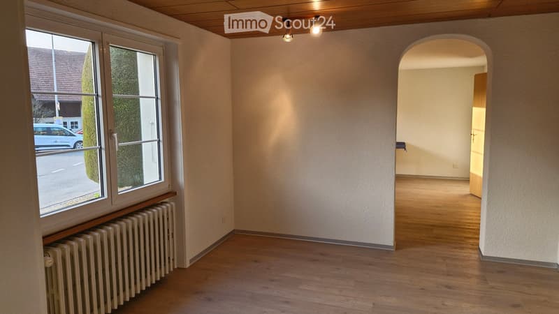 Empty room with wooden flooring, white walls, large window with view of street, radiator under window, and an arched doorway leading to another space.