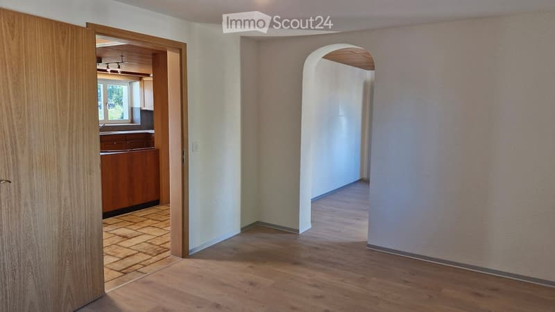 Empty hallway with wooden flooring, white walls, and an arched doorway leading to another room. A wooden door is open to reveal a kitchen area with cabinets and a window.