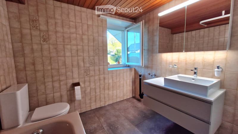 Modern bathroom with beige tiles, a white sink with a modern faucet, a toilet, a bathtub, and a large mirror above the sink. The room features a wooden ceiling and a window letting in natural light.