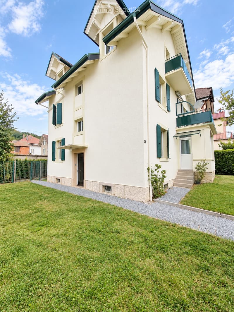Two-story house with light yellow walls, green shutters, and multiple balconies. The house features a well-maintained lawn and a paved pathway leading to the entrance.