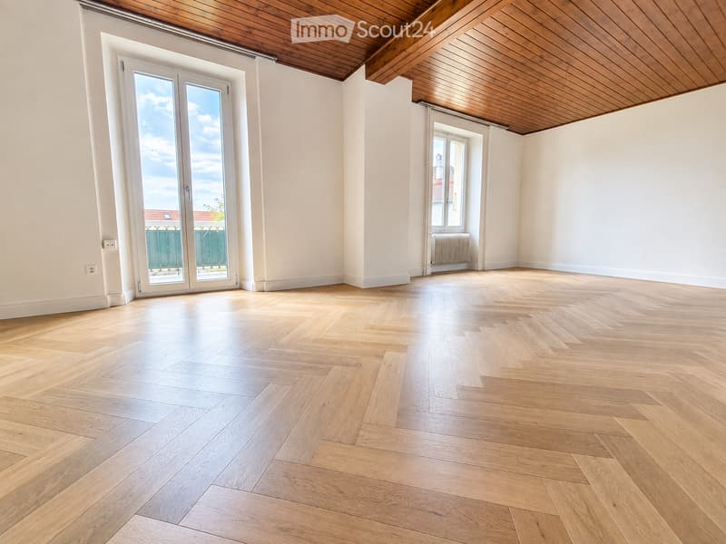 Empty room with light wooden herringbone flooring, white walls, and a wooden ceiling. Features include a large sliding glass door leading to a balcony and a smaller window with a radiator below.