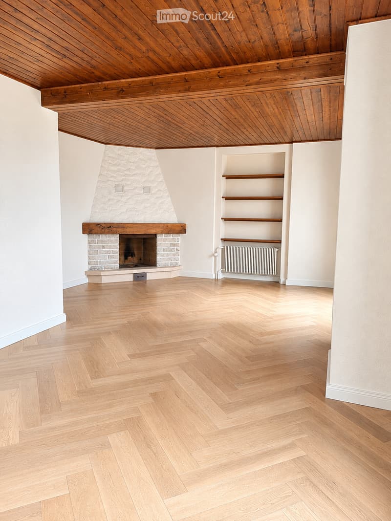 Empty room with wooden herringbone flooring, white walls, exposed wooden ceiling beams, and a stone fireplace with wooden mantel.