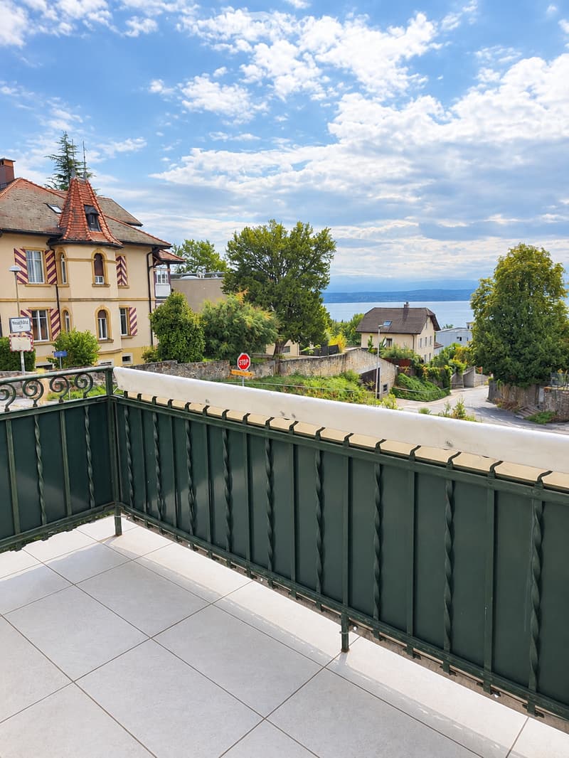 Balcony with green metal railing, tiled floor, overlooking a scenic view of a town, lake, and distant mountains.