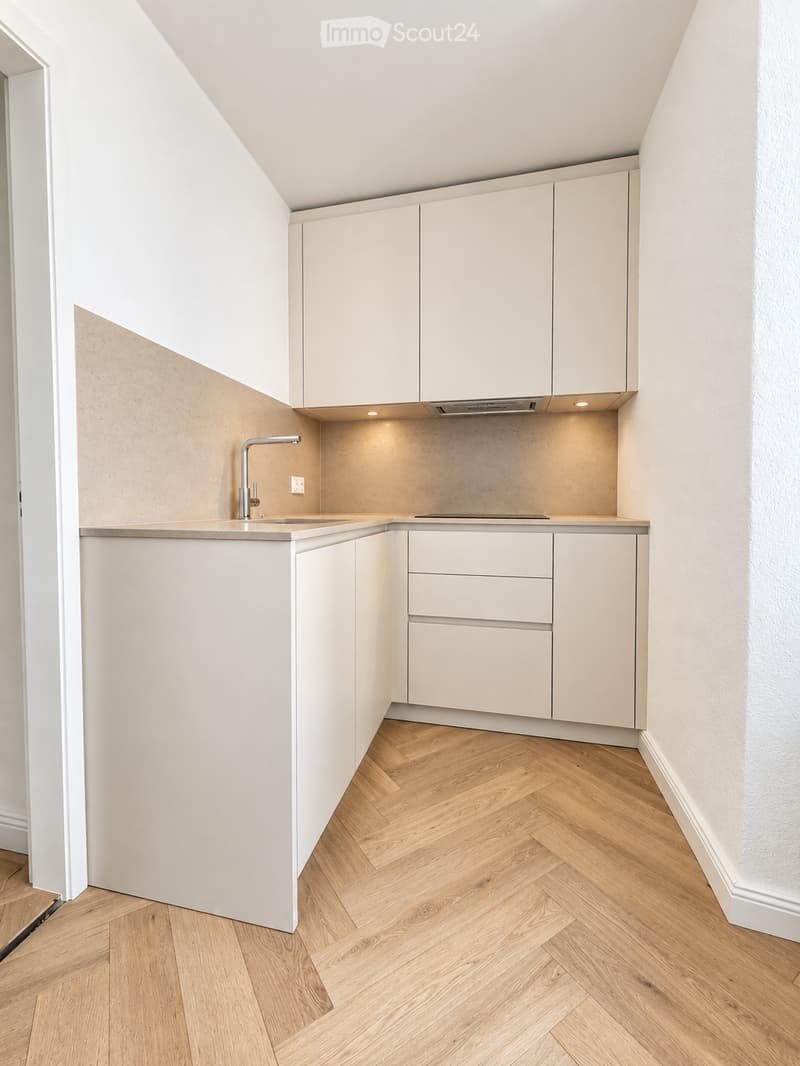 Modern, compact kitchen with light-colored cabinets, a sleek countertop, and a built-in sink with a modern faucet. The floor features herringbone-patterned wooden planks.