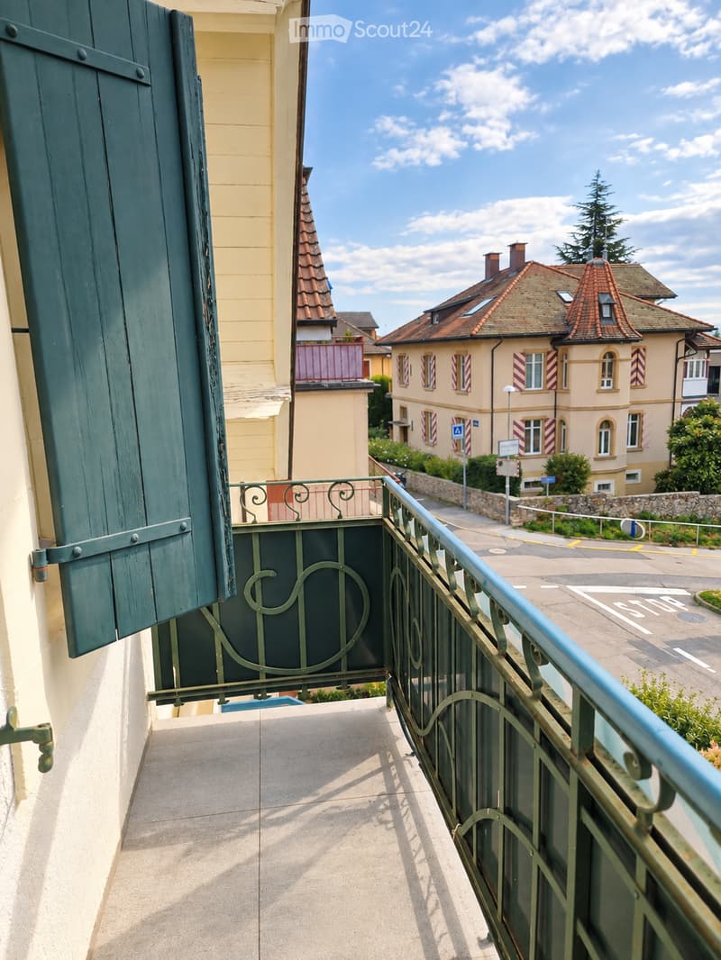 Balcony with green metal railing, overlooking a street lined with traditional European houses featuring pitched roofs and shutters.