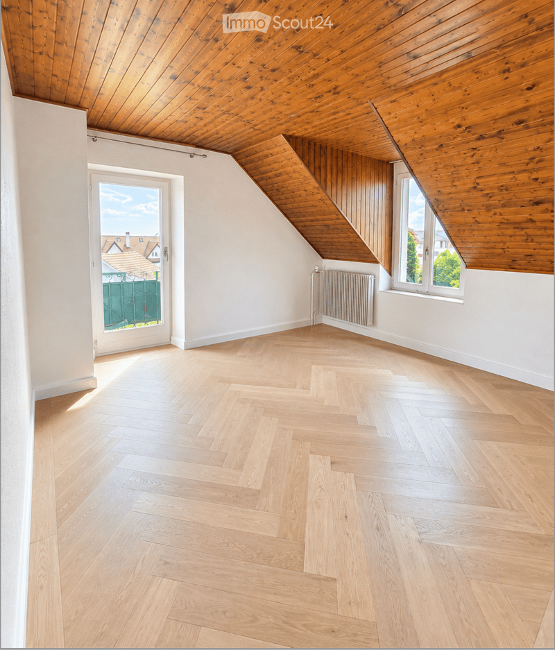 Empty attic room with wooden herringbone flooring, white walls, exposed wooden ceiling beams, and large windows offering natural light and a view of the neighborhood.