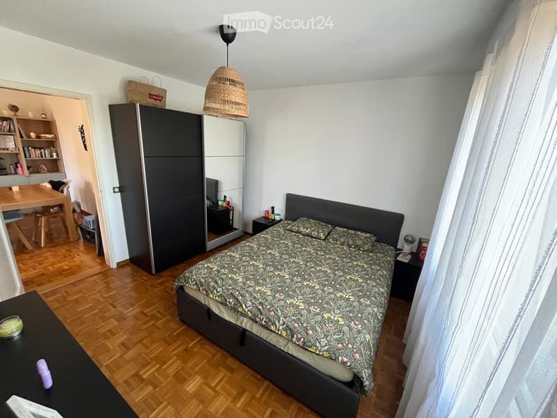 A furnished bedroom with a wooden floor, a large bed with a patterned duvet, a nightstand, a small table, a wardrobe, and a shelf in the adjacent area.