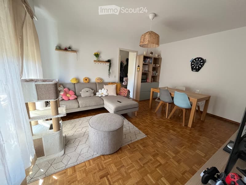 Living room with a grey sectional sofa, a round grey stool, a cat tree, a wooden dining table with four chairs, a bookshelf, and a large screen on a table.