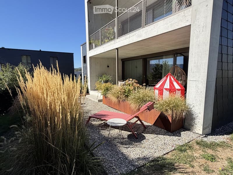 Modern outdoor area with a red lounge chair, small white table, wicker hanging basket, and tall grasses in planters near a contemporary building.