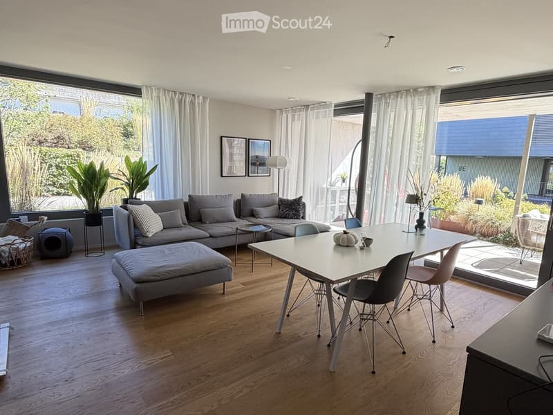 Modern living and dining area with wooden flooring, large windows, grey sofa, ottoman, coffee table, dining table with chairs, plants, and decorative items.