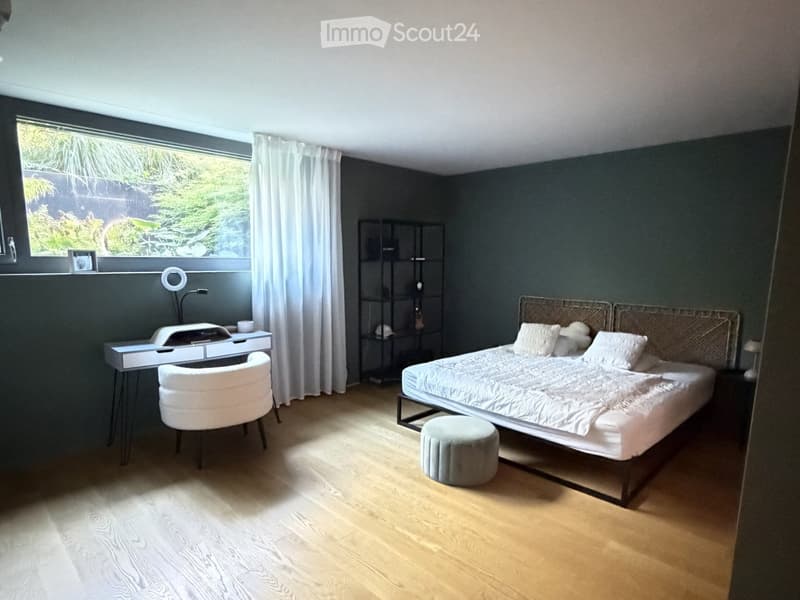 Modern bedroom with a large bed, white bedding, a round chair, a small stool, a black shelving unit, and a white desk with a lamp.
