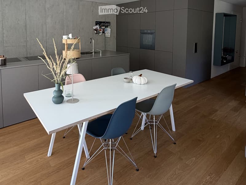 Modern kitchen with grey cabinets, white dining table, four chairs, vase with dried plants, lamp, and a sink area with faucet and oven.