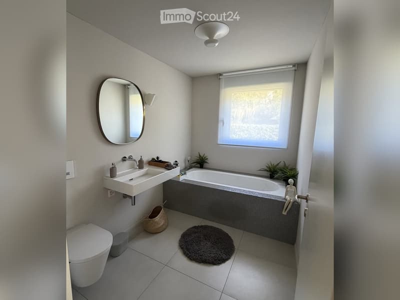 Modern bathroom with a white sink, bathtub, toilet, round mirror, and small decorative plants. Features a large window with blinds and a round rug on the floor.