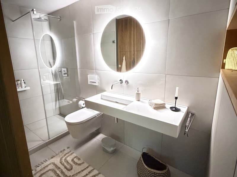 Modern bathroom with a white sink, toilet, glass shower enclosure, and large round mirror above the sink. Includes a towel, candle, and small basket on the floor.