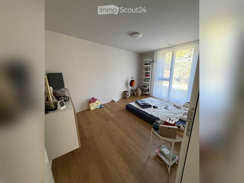 A well-lit bedroom with wooden flooring, a bed with a mattress and pillows, a small table with a screen, a shelf with books, and a toy on the floor.