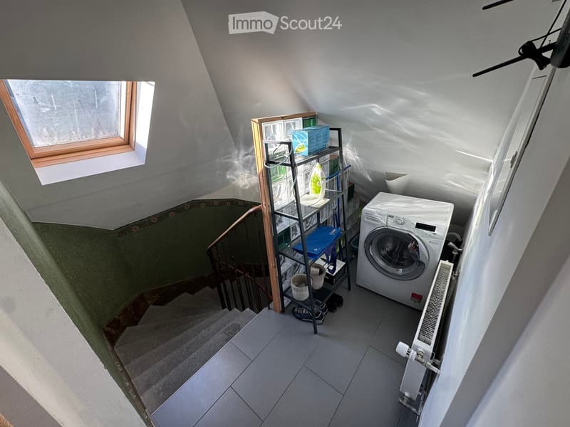 Small room with a washing machine, a metal shelf holding various items, and a staircase leading upwards.