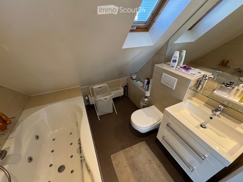 Modern bathroom with a bathtub, toilet, sink, mirror, and various toiletries. Features skylight windows and a small shelf with cups and cleaning supplies.