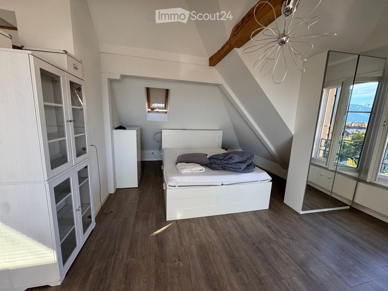 Bright attic bedroom with wooden flooring, white bed, wardrobe, large mirror, and a view through the window.
