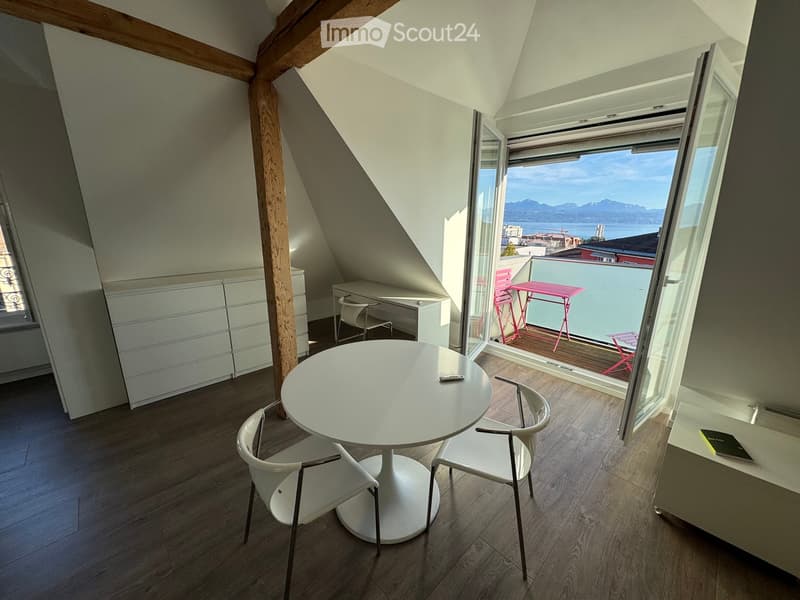 Bright attic room with wooden floors, white furniture, and a large window offering a scenic view of the lake and mountains. The room features a round white table with two chairs, a desk with a chair, and a white cabinet.