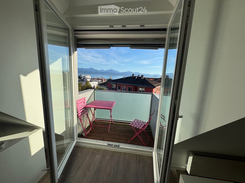 Balcony with wooden floor, pink table and chairs, overlooking a scenic view of a city and mountains.