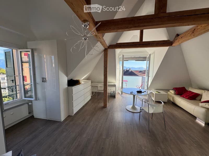 Modern attic living space with wooden beams, exposed ceiling, large windows offering a scenic view, and contemporary furniture including a sofa, dining table, chairs, and a cabinet.
