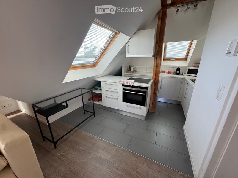 A modern, compact kitchen area with white cabinets, a built-in oven, microwave, and a small black table with a glass top. The space features wooden flooring and grey tiled flooring in the kitchen area, with skylight windows providing natural light.