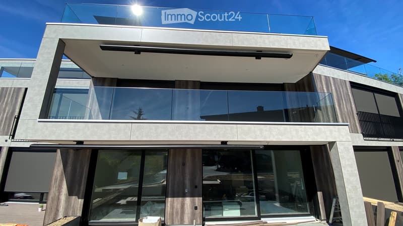 Modern two-story house with large glass windows and balconies, featuring a sleek concrete and wood exterior design.