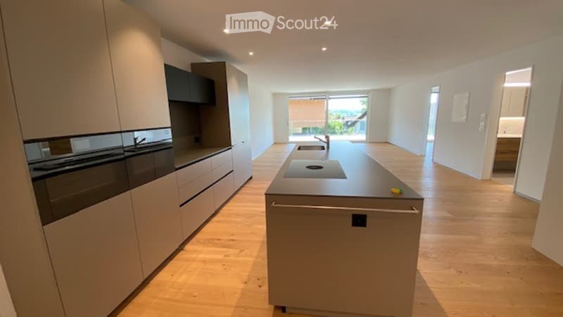 Modern kitchen with light wood flooring, built-in appliances, and a central island with a sink and faucet. Large windows provide a view of the outdoor area.