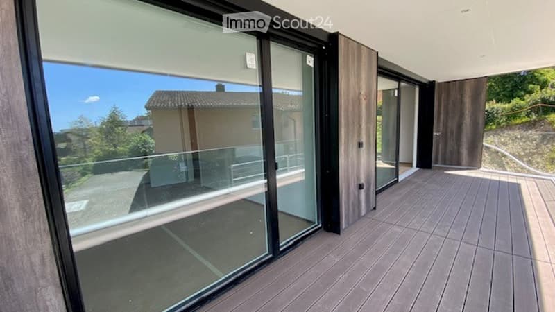 Modern balcony with large glass sliding doors, wooden decking, and scenic view of surrounding landscape and buildings.