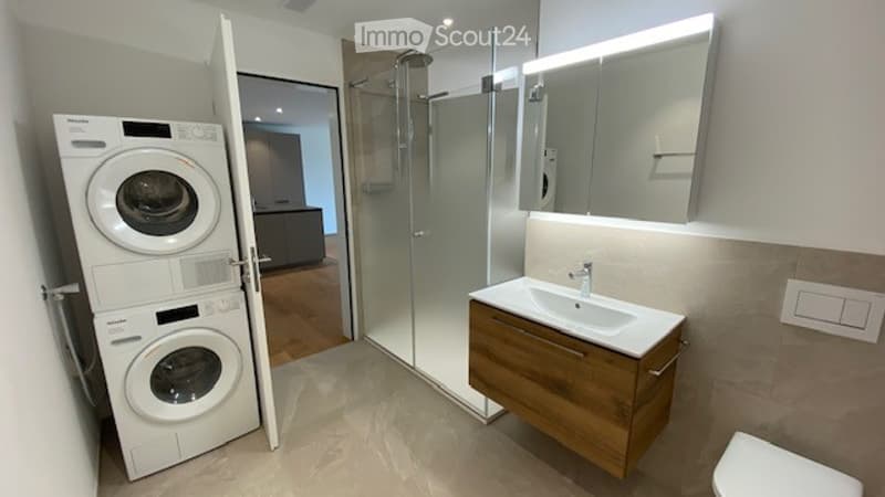 Modern bathroom with a glass-enclosed shower, a wooden vanity with a sink, a large mirror, and a stacked washer/dryer unit.