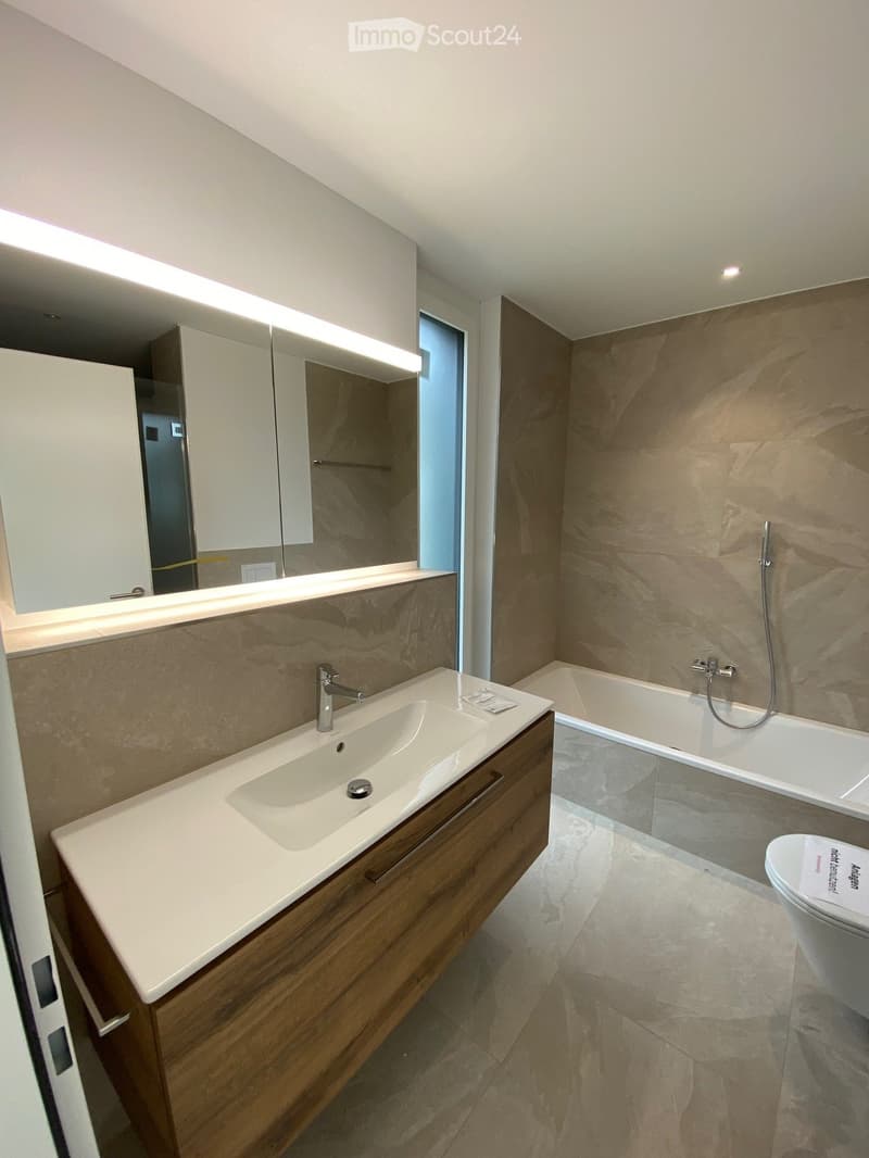 Modern bathroom with a double sink, wooden vanity, large mirror, bathtub, and toilet. The walls are tiled in a beige marble pattern, and the floor is also tiled in a similar style.