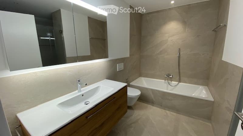 Modern bathroom with a white sink, wooden cabinet, large mirror, bathtub, and toilet. The walls and floor are tiled in a light beige color, and the lighting is bright and contemporary.