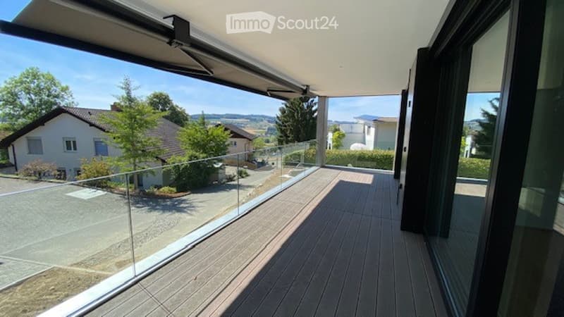 Modern balcony with wooden decking, glass railing, and scenic view of surrounding houses and landscape.