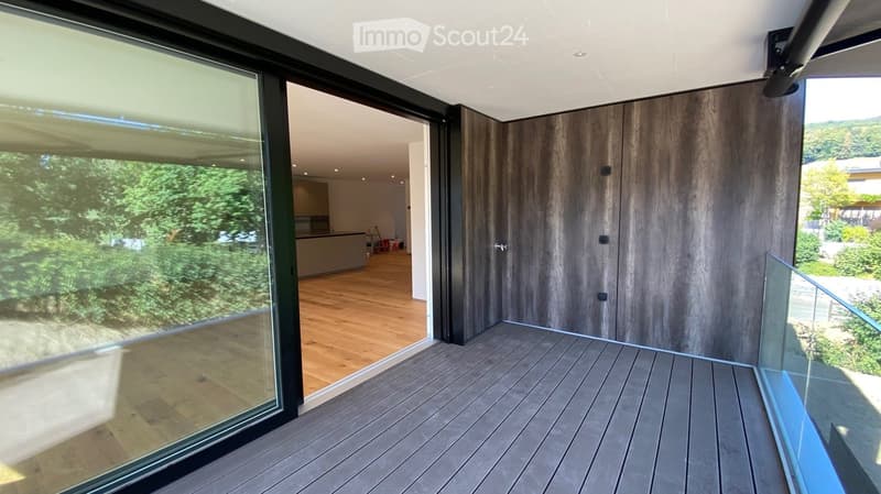 Modern balcony with wooden decking, large glass sliding doors, and a view of greenery.