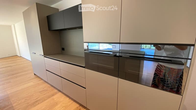Modern kitchen with white and grey cabinets, a central island, and a built-in oven. The floor is light wood, and the space is well-lit with natural light from a large window.