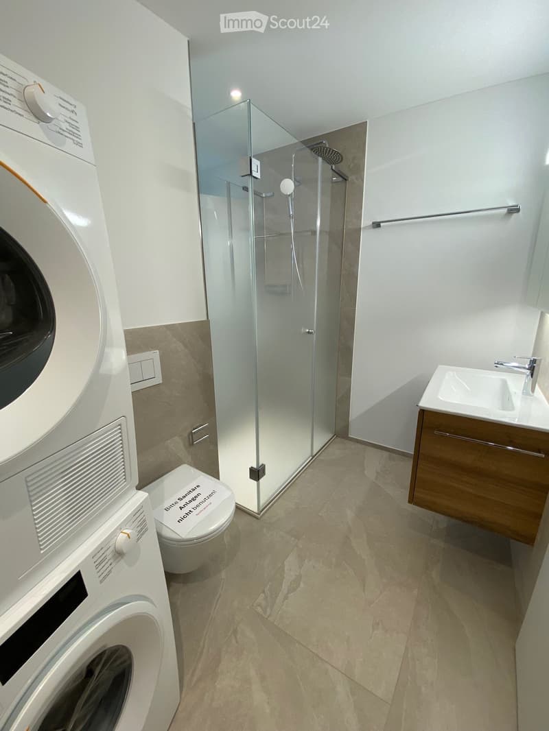 Modern bathroom with a glass-enclosed shower, a toilet, a sink with a wooden cabinet, and a washing machine.