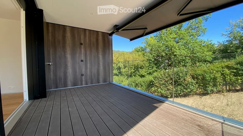 Modern wooden deck with glass railing, overlooking a lush green landscape with trees.