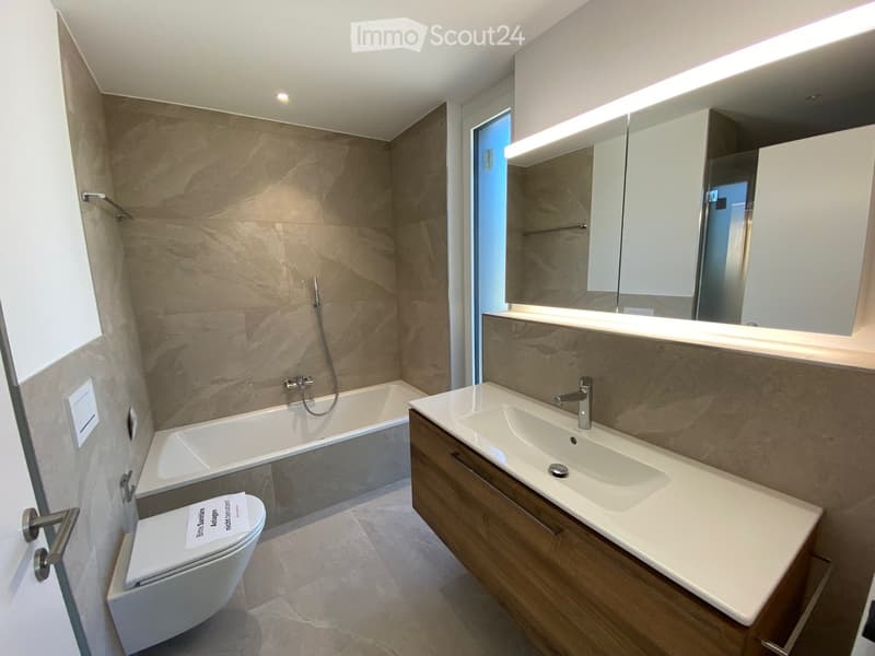 Modern bathroom with a bathtub, sink, toilet, and large mirror with built-in lighting. The walls are tiled in a beige marble pattern, and the floor is tiled in a light beige color.