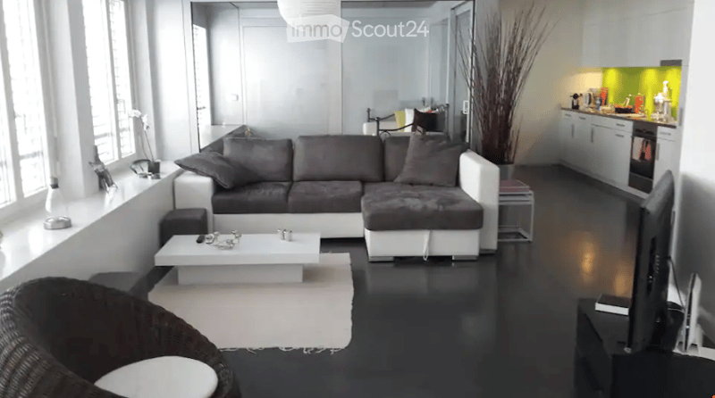 Modern living room with a grey sectional sofa, white coffee table, black wicker chair, and a flat-screen TV. The adjacent kitchen features white cabinets, a stainless steel oven, and a modern faucet.
