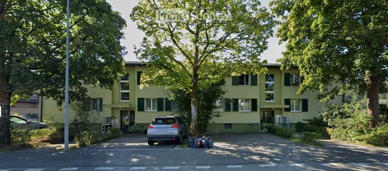 A two-story residential building with light yellow walls, green shutters, and multiple windows. The building is surrounded by trees and has a driveway with a parked car and some bags.