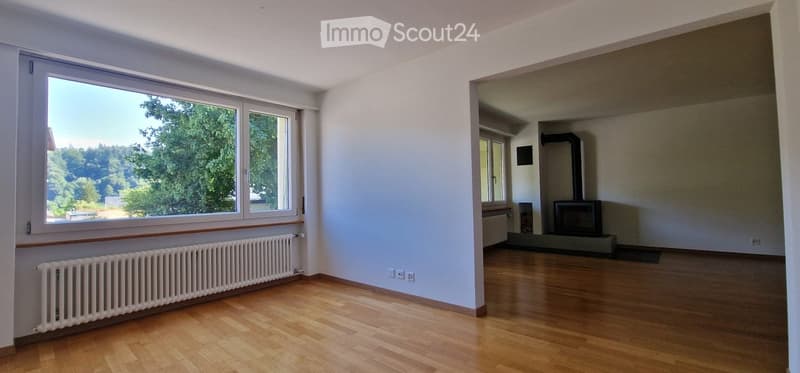 Empty room with wooden flooring, white walls, large windows with a view of greenery, radiator, and a fireplace in the adjacent room.