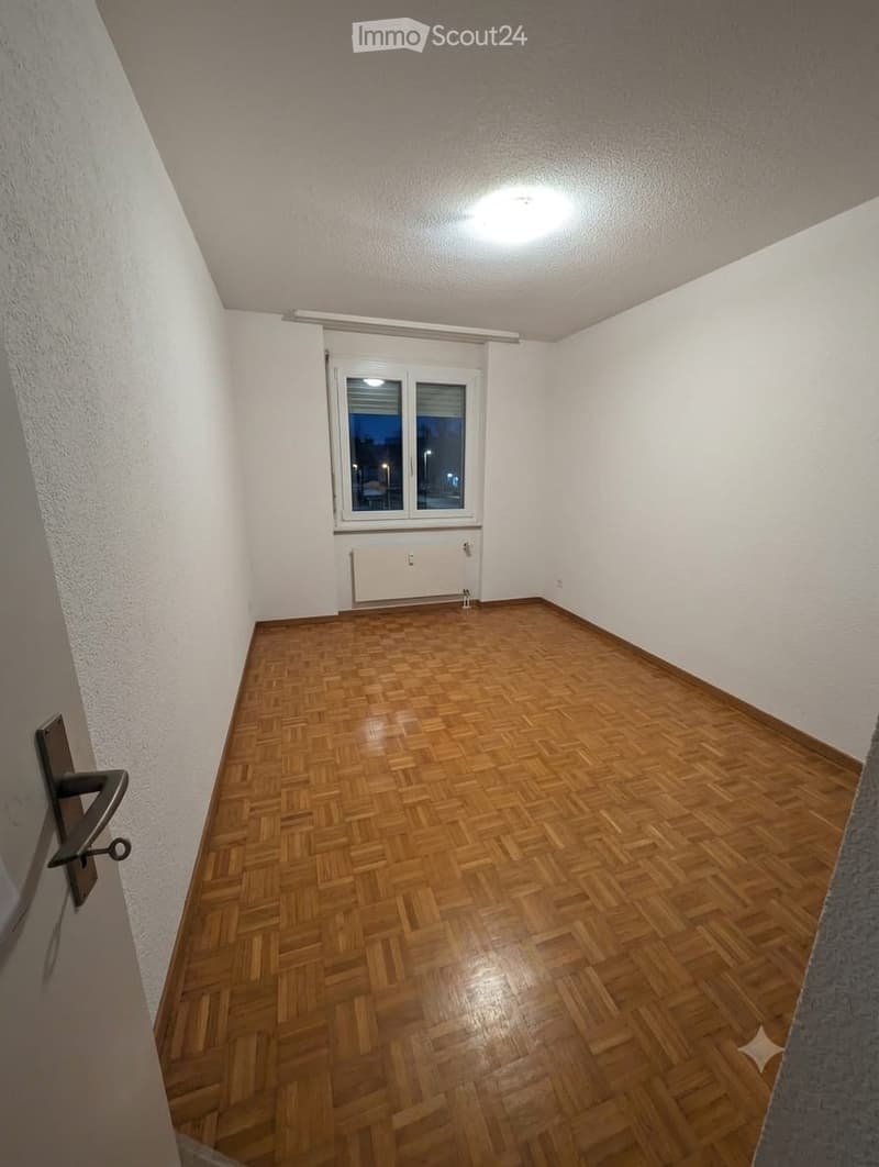 Empty room with wooden parquet flooring, white walls, and a single window with blinds. No furniture or equipment is present.