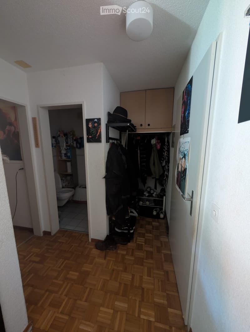 A narrow hallway with wooden flooring, white walls, and a ceiling light fixture. A closet with hanging clothes and shoes is visible, along with an open door leading to a bathroom.